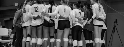 Black and white photo of the women's volleyball team standing on the court with their arms around eachother in a huddle.
