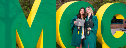 Two female MCC students stand in their graduation gowns holding their diplomas in front of large MCC letters outside after commencement May 2, 2025.