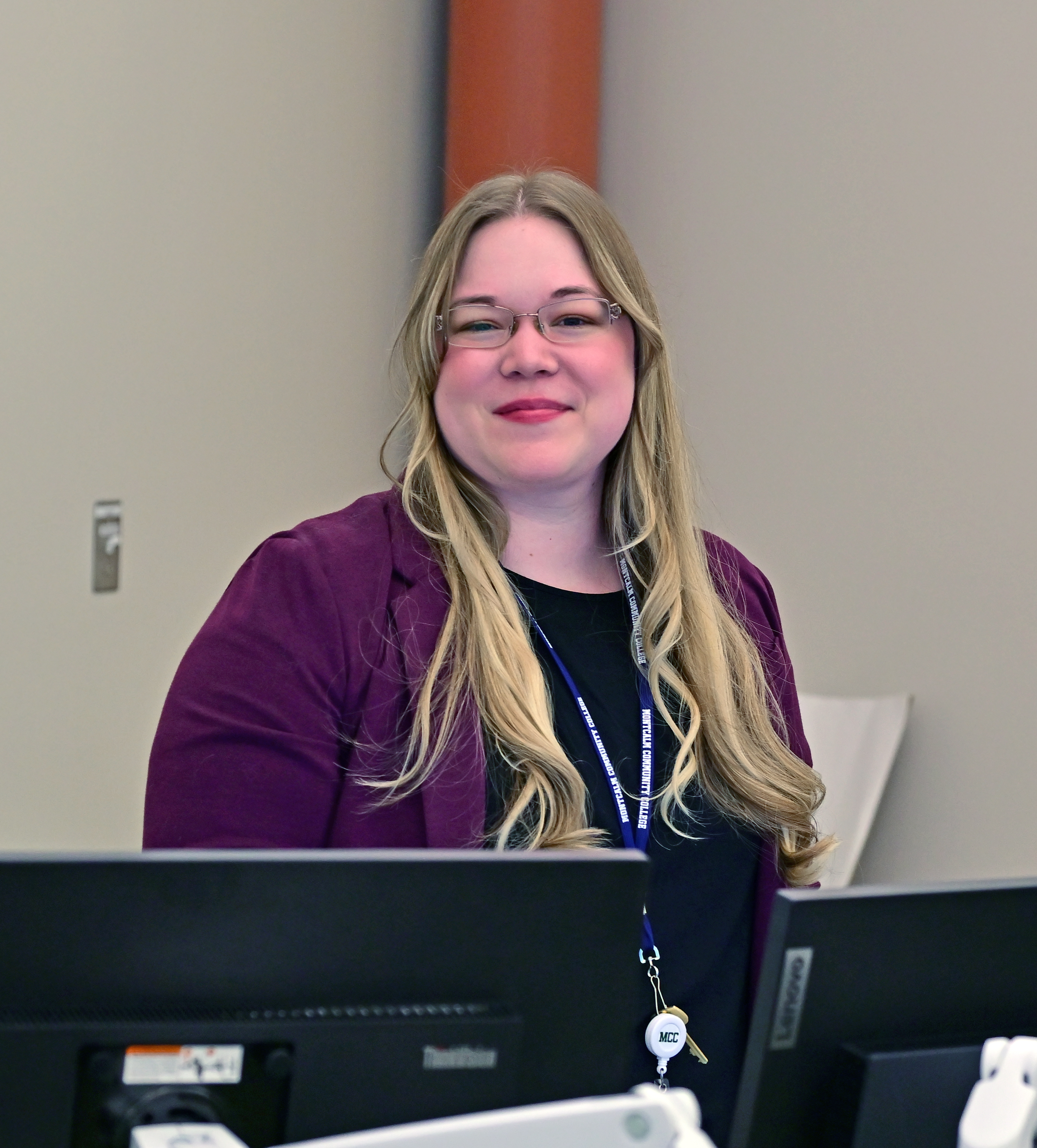 English Instructor, Devan Bridget wearing a purple blazer and standing in front of computer screens smiling.