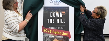 Two women revealing a poster with the cover of the book "Down the Hill" by Susan Hendriks on it during the One Book One County Montcalm 2025 kickoff event.