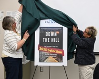 Two women revealing a poster with the cover of the book "Down the Hill" by Susan Hendriks on it during the One Book One County Montcalm 2025 kickoff event.