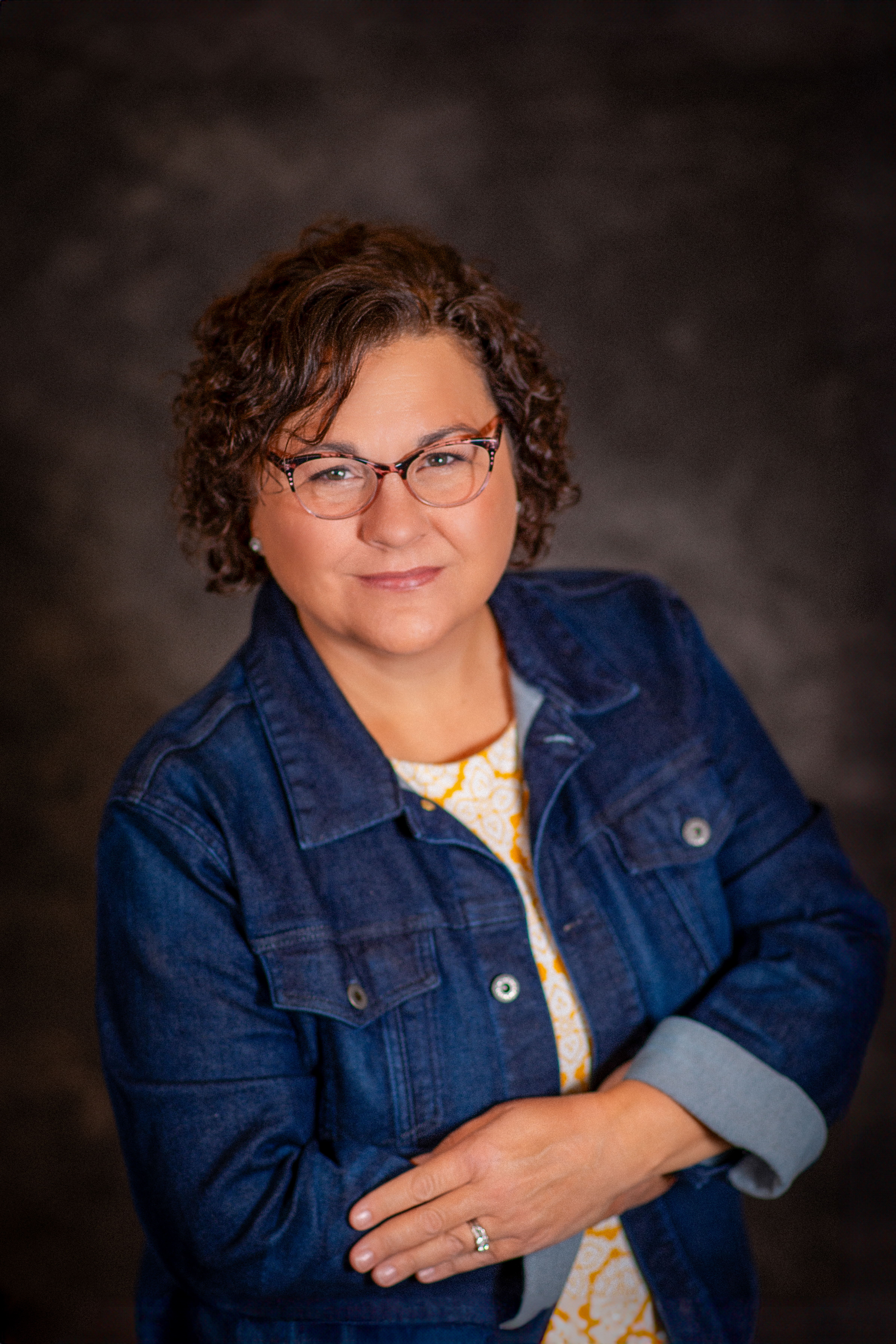 MCC Faculty Member Krsiten Diehl smiling and wearing a jean jacket, orange and white patterened shirt and glasses.