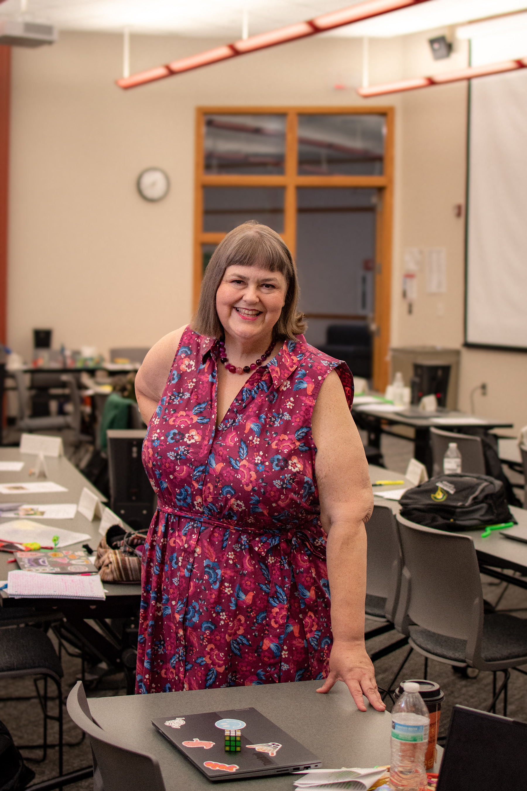 English Instructor Greta Skogseth in a pink floral dress and short brown hair standing in front of a desk with classroom behind her.