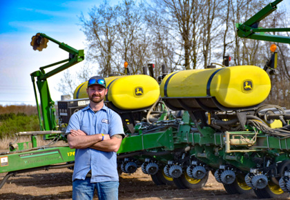 Man with brown beard and mustache in blue work shirt and jeans crosses arms and smiles at camera. Man is wearing a ball cap with sunglasses on top and is posing in front of John Deere tractor equipment.