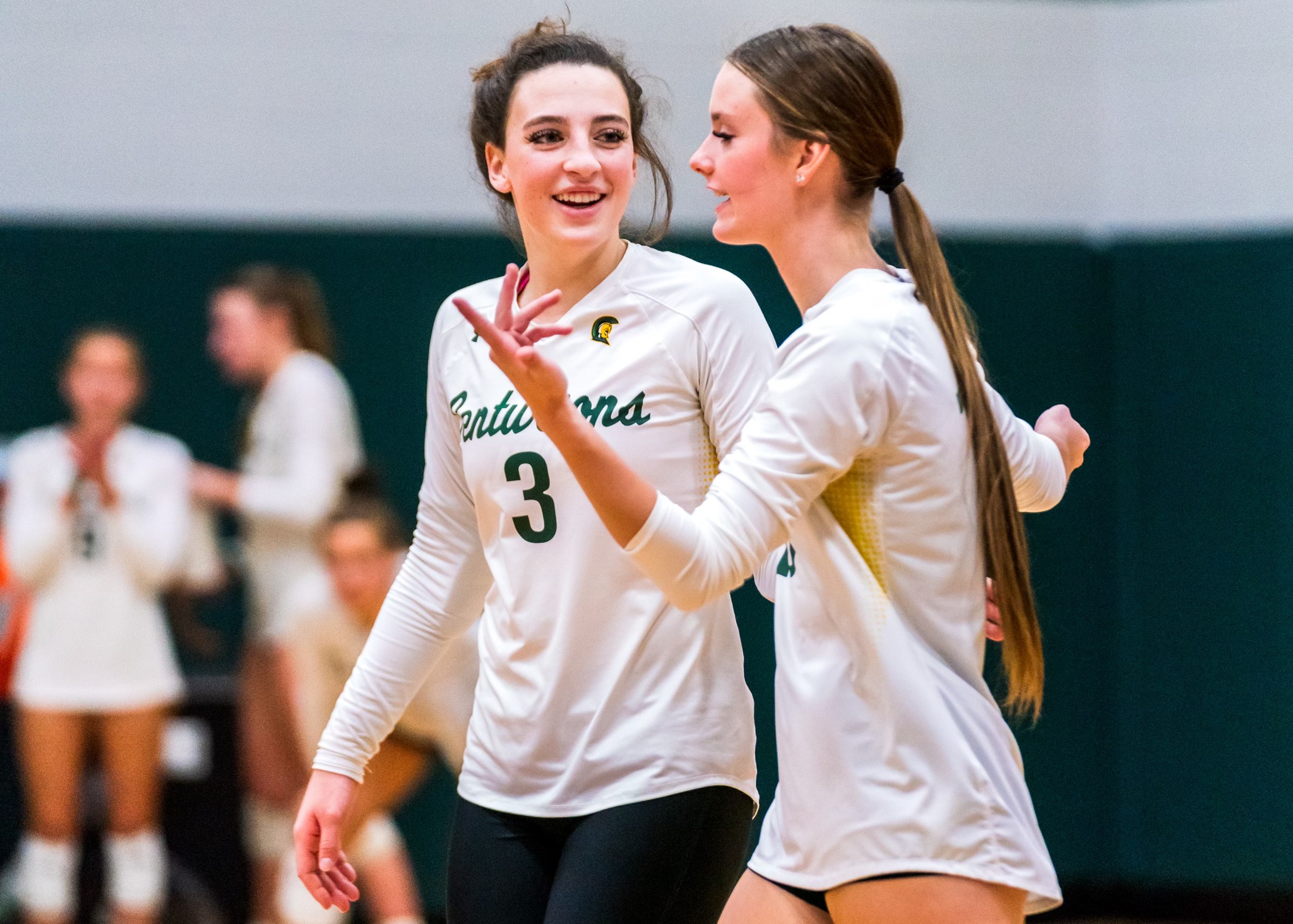 Two MCC Volleyball players talk on the court during a match with smiles on their faces.