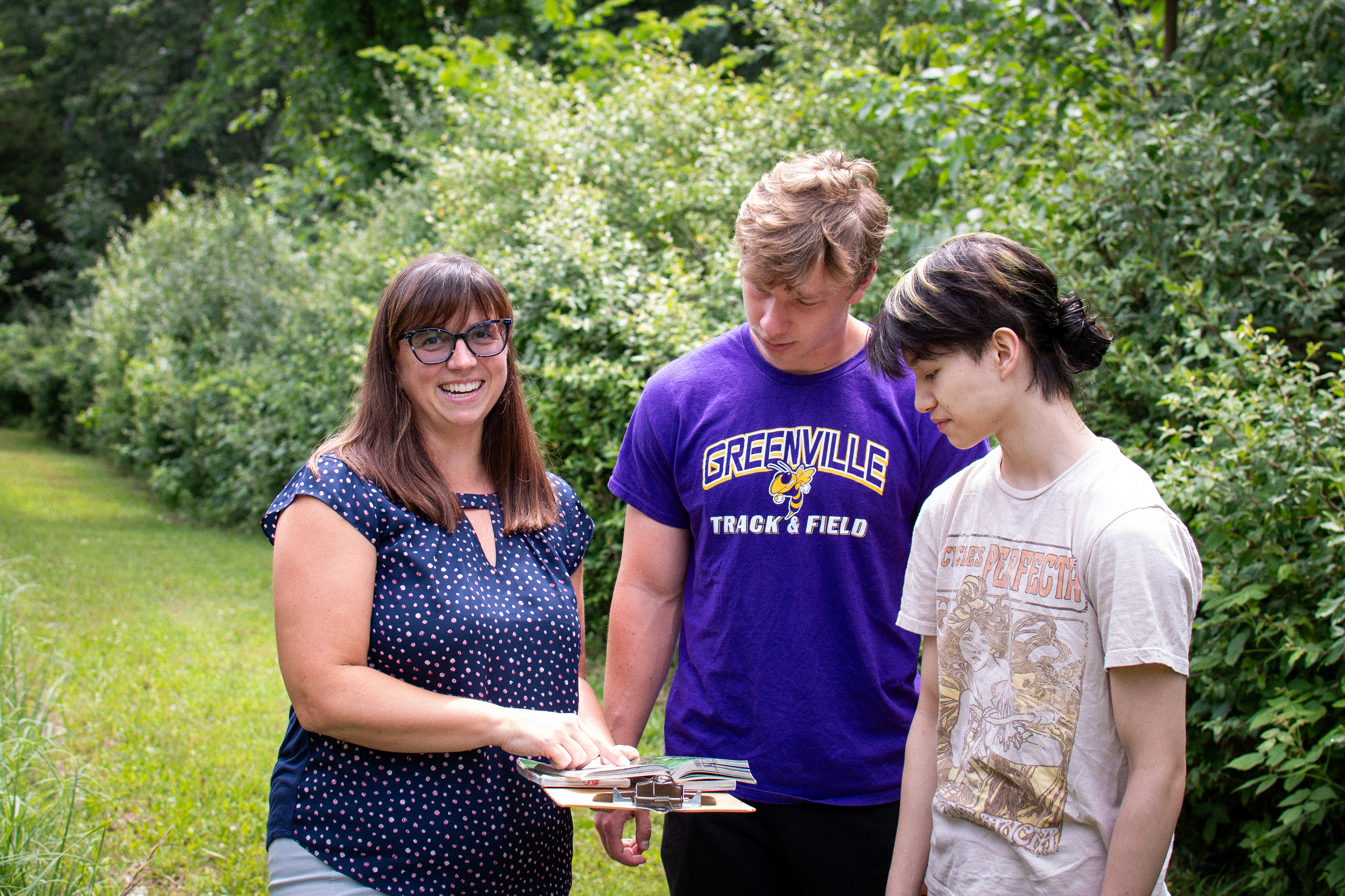 Instructor, Heather Wesp with medium length brown hair and glasses wearing a navy blue shirt with white dots and showing two students a clipboard outside with foliage in the background.