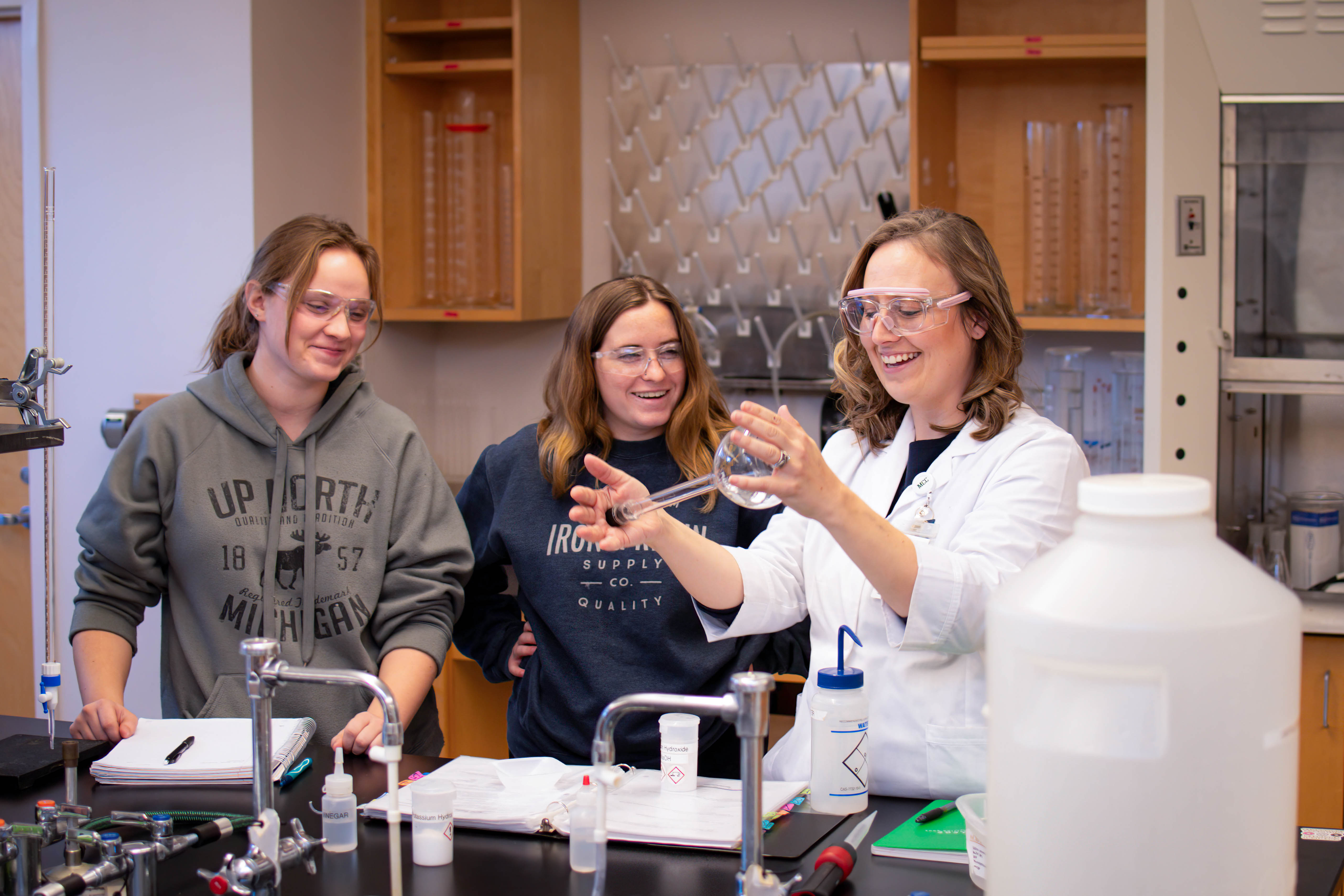 Instructor Sara Rittersdorf in the Chemistry lab holding a beaker with two students.