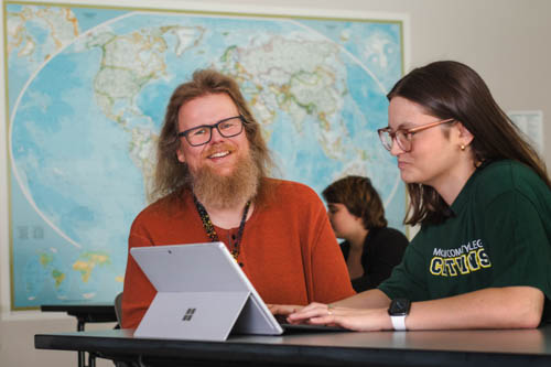 Counselor, Tore Skogseth, with light brown hair, a beard and glasses wearing an orange shirt sitting at a desk looking at a tablet with a student with brown hair, glasses and wearing a green Centurion t-shirt.